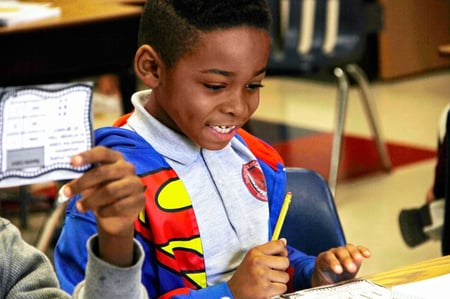 A young student holds a pencil and smiles down at the desk.