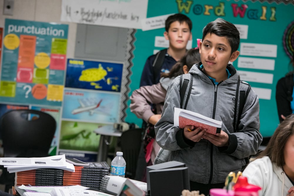 A child stands in a classroom holding a notebook. 