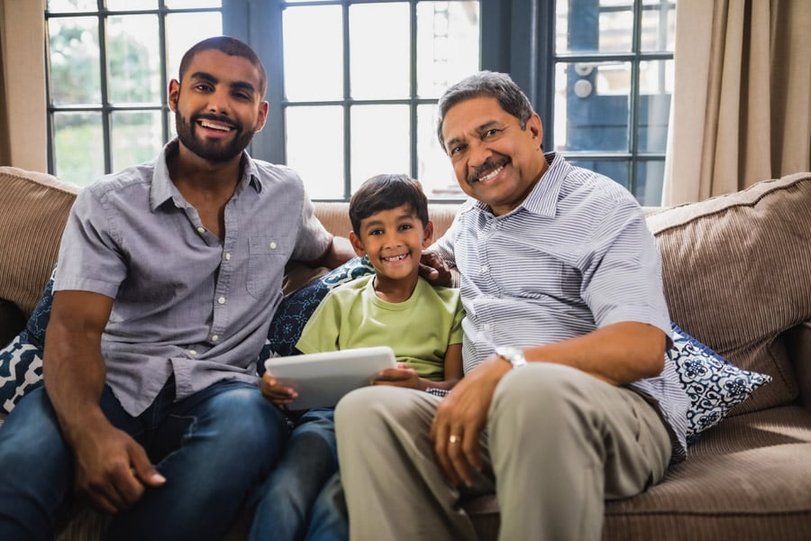 A young boy, father, and grandfather all sit on a sofa and smile at the camera.