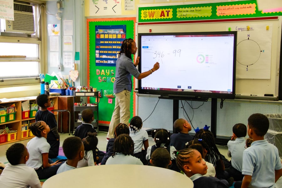 A male teacher writes on an electronic whiteboard while a class of kindergarten students watch.