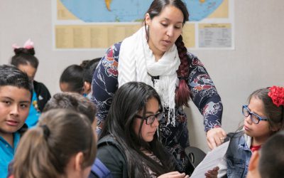 A teacher points to a paper that a student holds while seated at her desk.