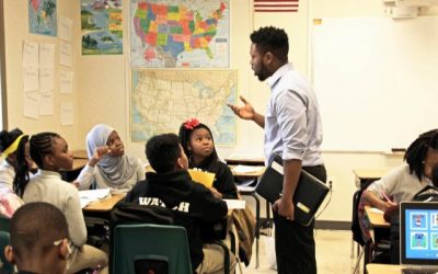 A male teacher addresses a group of four students seated at a cluster of desks. 