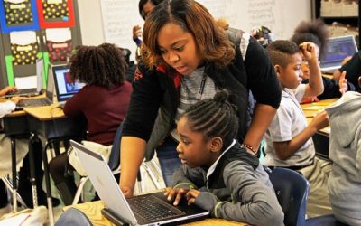 A female teacher leans over to point out something on the screen of a student's laptop. 